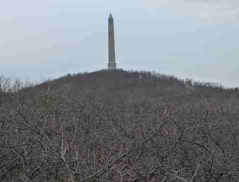 A tall stone obelisk monument stands atop a tree-covered hill under a cloudy sky, with bare branches in the foreground—reminiscent of views along the AT Section Hike near Mohican Outdoor Center. No leaves are visible on the trees.