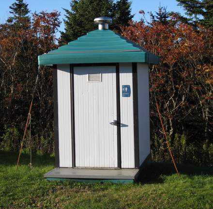 A small, white outdoor restroom with a green roof stands on grass, surrounded by bushes and trees. A blue sign on the door indicates it is a public toilet, supporting Leave No Trace principles and responsible outdoor ethics.