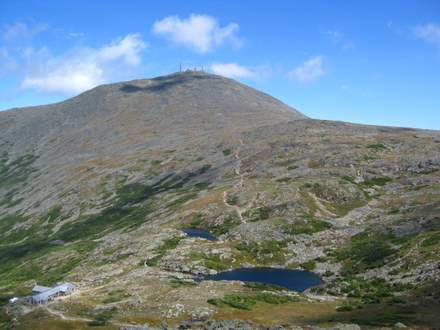 A rocky, grassy mountain landscape features small ponds and a clear path leading up to a summit. Near the bottom left, a building sits close to the Edmands Path. The sky above is bright blue with a few clouds.