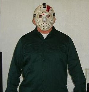 A person wearing a white hockey mask with black and red details and a dark green shirt stands against a plain light-colored wall, looking as if they're ready to discuss sleeping bag temperature ratings for your next camping trip.