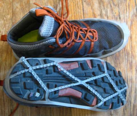 A pair of dark sneakers with orange laces, one upright and the other flipped to show the sole. The sole features a Yaktrax crisscross traction device, and both shoes are on a wooden floor.