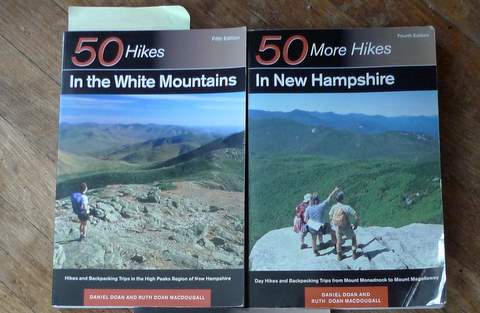 Two hiking guidebooks from the 50 Hikes Series by Dan Doan are shown side by side on a wooden surface, each featuring hikers on scenic mountain trails: 50 Hikes in the White Mountains and 50 More Hikes in New Hampshire.