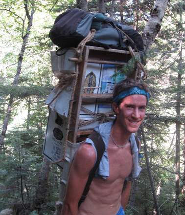 A smiling, shirtless hiker known as HutMan, wearing a headband, carries a large backpack—complete with bookshelf and other items—through a sunlit forest, perhaps fueled by an adventurous Diet Plan perfected for AMC trail journeys.