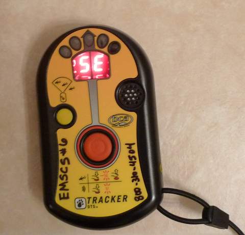 A yellow and black digital avalanche beacon labeled “TRACKER DTS” features a red button, a screen displaying SE, handwritten text EMSCS #6 Hash-mic 08, and comes with a wrist strap attached.