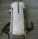 A lightweight, white Porter Pack by Hyperlight Mountain Gear with black straps and a roll-top closure sits on a wooden deck. The backpack has minimal padding and an adjustable hip belt, perfect for your next expedition.