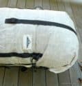 A white Hyperlight Mountain Gear 4400 Porter Pack with black straps lies on a wooden surface. This ultralight expedition backpack features a rectangular logo with a mountain and the word “HMG.”.