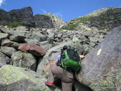 A person with a green backpack climbs over large, rocky boulders on the Boot Spur Loop of Mt Washington, following a yellow trail marker painted on a rock under a blue sky.