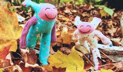 Two colorful, smiling plush toys with long arms and legs—creations by Trail Magic Dollmaker Arlette Laan—stand among autumn leaves outdoors. One toy is blue and pink; the other is white with pink accents, showcasing charming fiber arts.