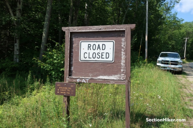 A wooden sign reads ROAD CLOSED next to an overgrown road with a parked white pickup truck. Trees and grass surround the area, while a small brown sign displays hiking trail info—almost as if this page does not exist anymore.