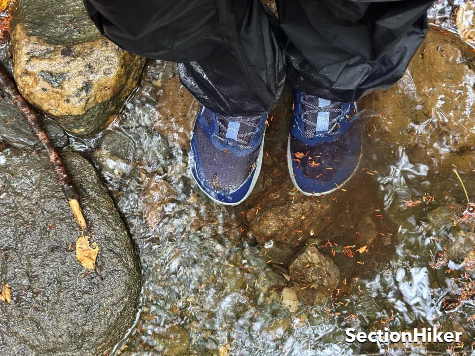 Close-up of a person wearing waterproof trail running shoes and black pants, standing in a shallow stream with rocks and leaves, with “SectionHiker” text in the bottom right corner.