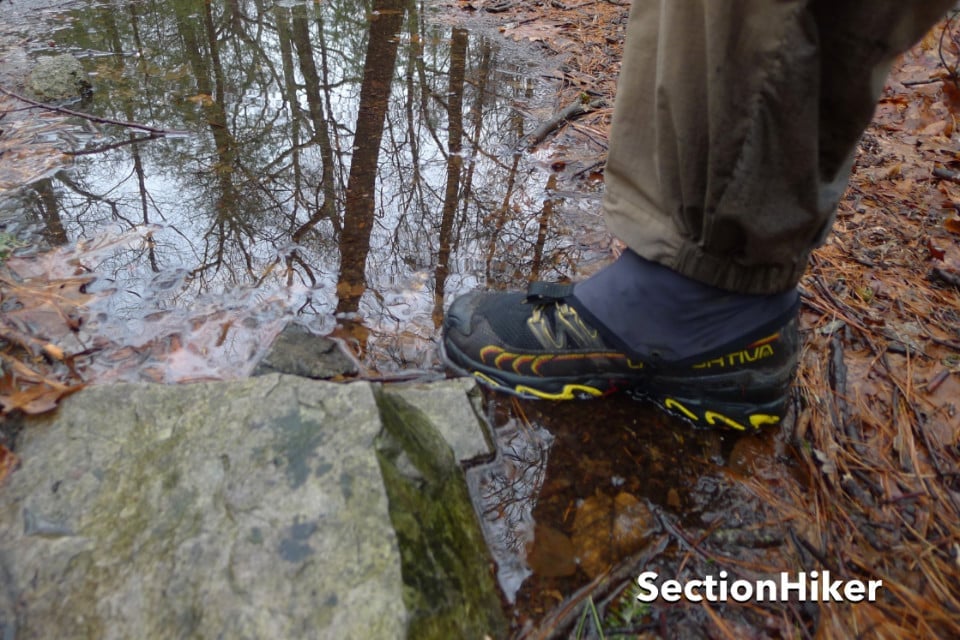 A person wearing waterproof trail running shoes and gaiters steps in a shallow puddle on a muddy, pine needle-covered forest path. The reflection of leafless trees appears in the water. SectionHiker is written at the bottom right.